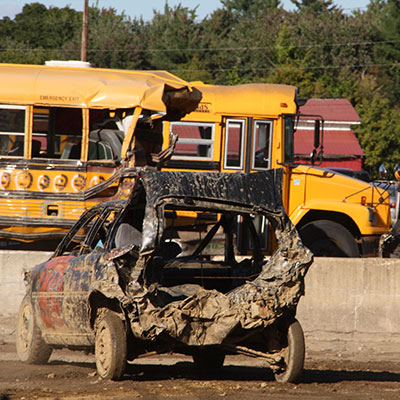 A muddy crunched up derby car is shown in front of two school buses one of which is also crushed